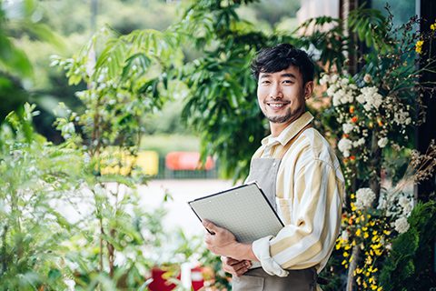 Portrait of confident young Asian male florist, owner of small business flower shop, standing in front of flower shop with clipboard on hand. He is smiling and looking at camera.